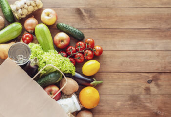 Paper grocery bag with fresh vegetables, fruits, milk and canned goods on wooden backdrop. Food delivery, shopping, donation concept. Healthy food background. Flat lay, copy space.